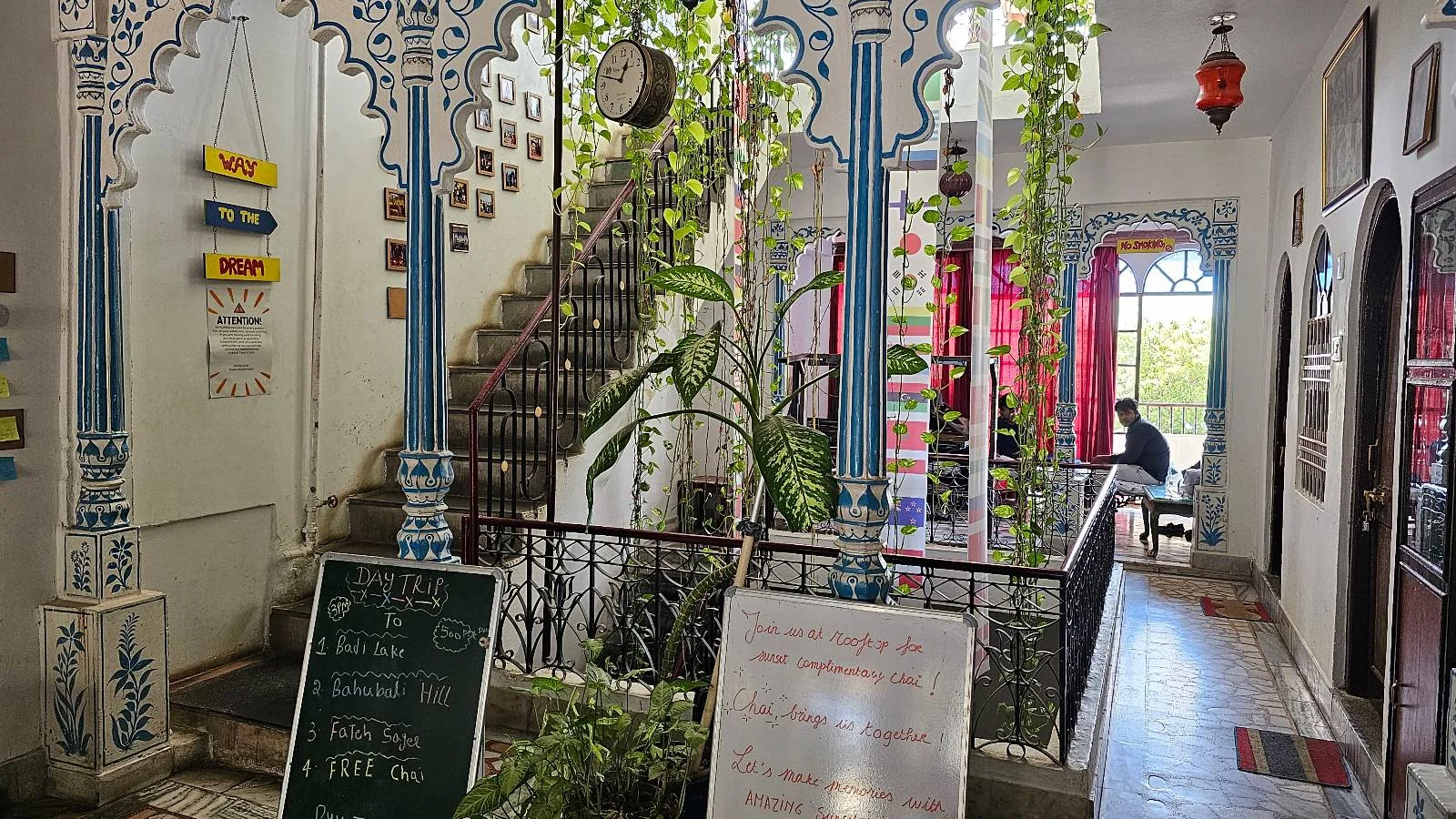 Colorful, ornate interior of a café or guesthouse with blue and white arches, hanging plants, chalkboard menus, and a staircase. Sunlight streams through windows, and a person sits at a table in the background.