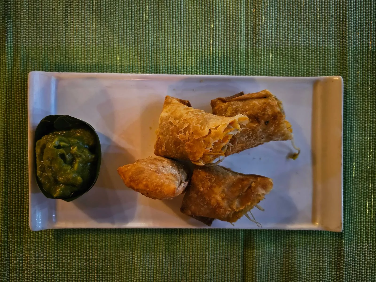 Four crispy spring rolls are arranged on a rectangular white plate with a small dish of green dipping sauce on the side. The plate is set on a woven green mat.