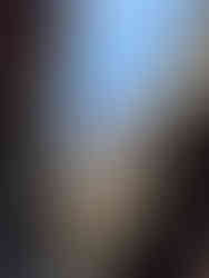 Stone faces carved into the ancient towers of a temple rise against a clear blue sky, viewed from a narrow passage with detailed stone walls and lush green plants growing out of the rocks.