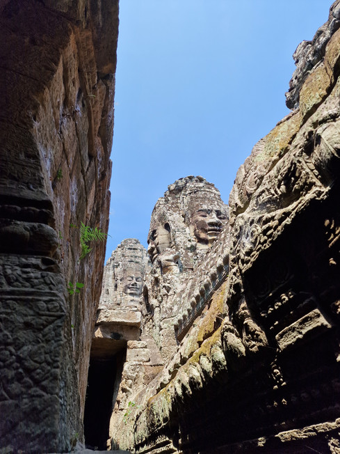 Stone faces carved into the ancient towers of a temple rise against a clear blue sky, viewed from a narrow passage with detailed stone walls and lush green plants growing out of the rocks.