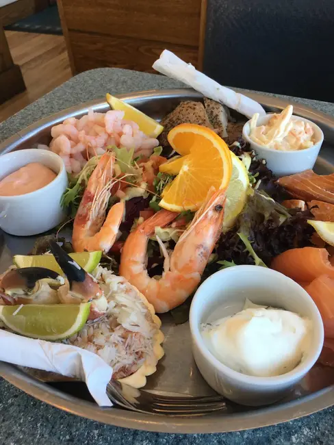 A seafood platter featuring prawns, a scallop with lime, and assorted greens. Accompanied by orange slices, marinated vegetables, and dipping sauces in small bowls.