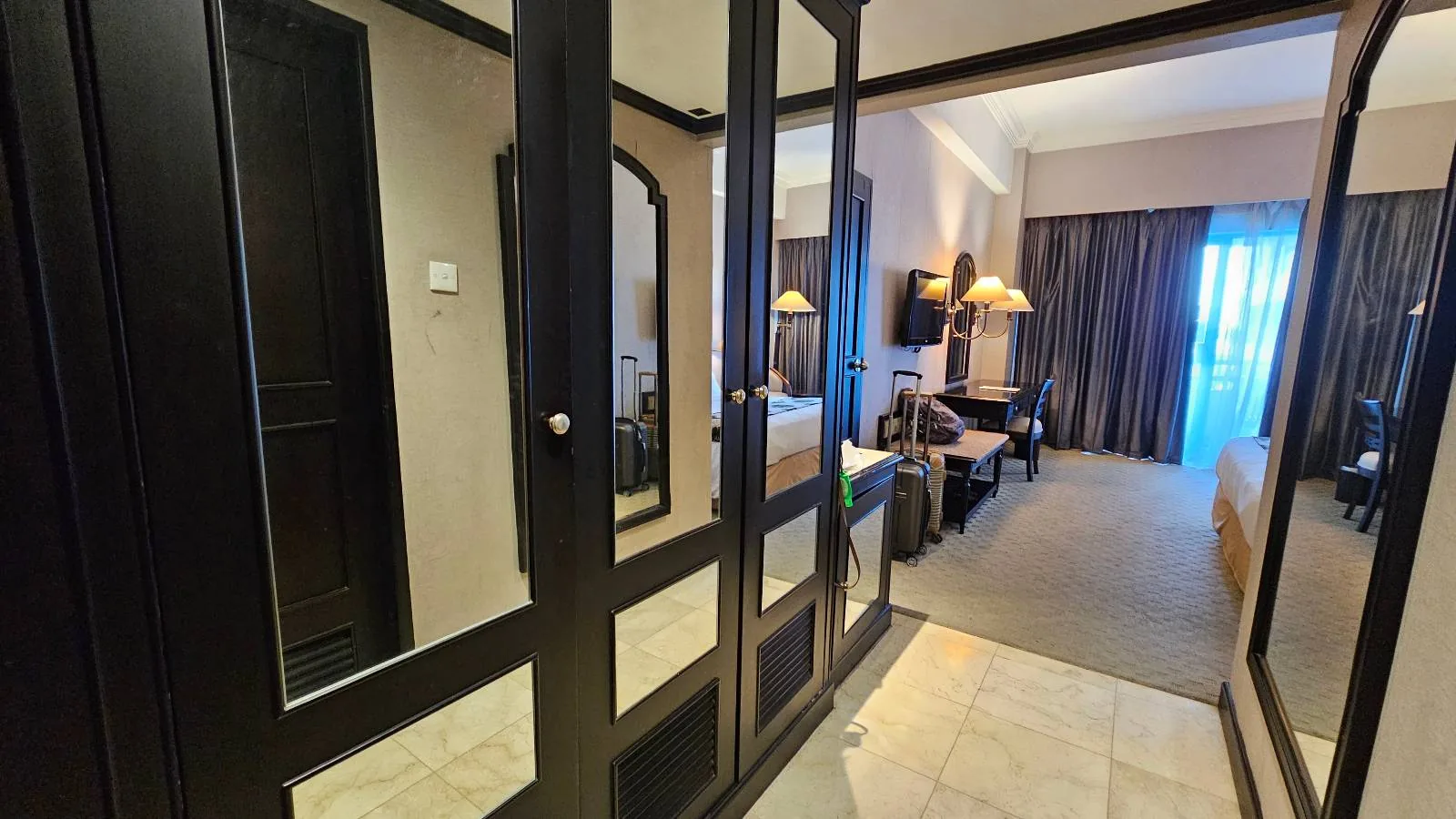 Hotel room interior with a view from the entrance, showing a mirrored closet on the left. The room has a bed, lamps, a desk, and a TV. Natural light filters through large windows covered with dark curtains in the background.