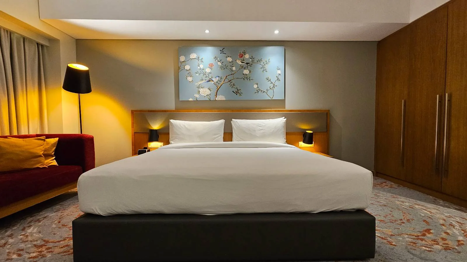 A modern hotel room with a large bed, white bedding, two bedside tables with lamps, a red sofa, a standing lamp, and a wall painting above the bed. Warm lighting creates a cozy atmosphere.