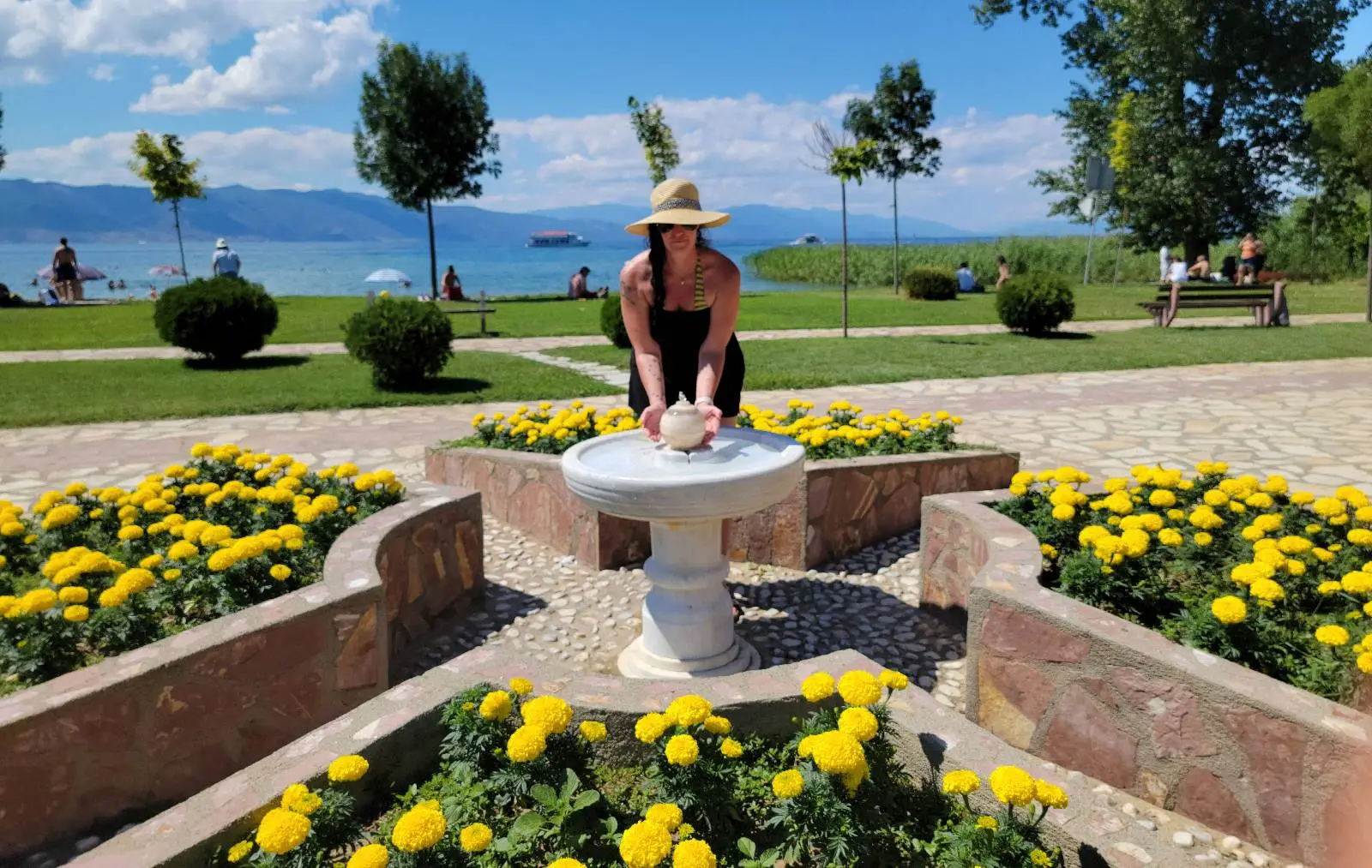 A person in a straw hat leans over a small stone table surrounded by vibrant yellow flowers in a park. A scenic lake and several trees are in the background under a blue sky with fluffy clouds.