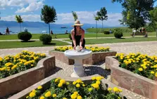 A person in a hat leans over a stone table in a garden filled with flowers in their favorite colors. A scenic backdrop of a lake and mountains is visible under the clear blue sky, creating the perfect setting for capturing photos.