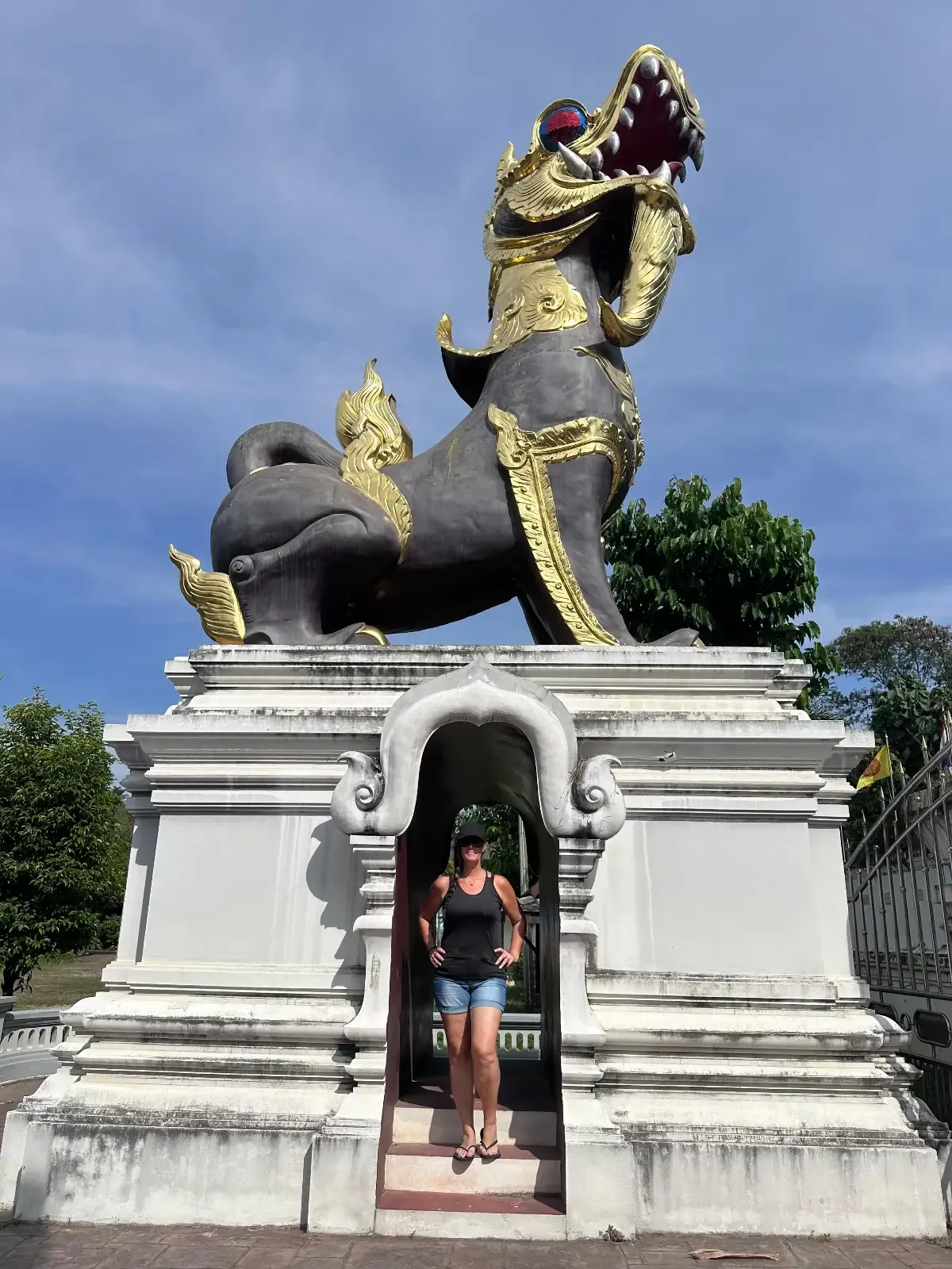 A person stands beneath a large, ornate statue of a mythical lion-like creature with gold decorations, set on a white pedestal outdoors under a blue sky.
