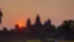 Sunset over Angkor Wat with orange sky, silhouetted temple towers, and palm trees. Tourists stroll in the foreground. Tranquil mood.