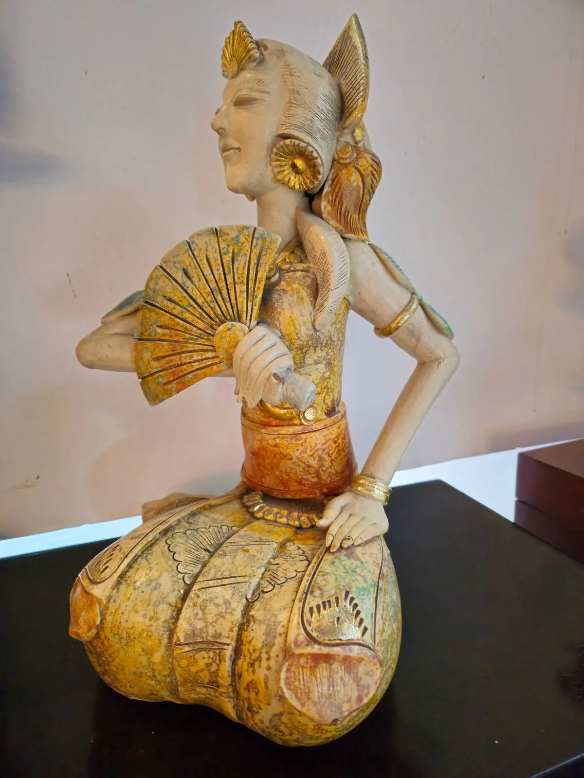 A detailed sculpture of a mythical woman with fox-like ears, holding a fan. She is dressed in traditional attire with intricate patterns, and is posed gracefully. The sculpture has a warm, earthy color palette.