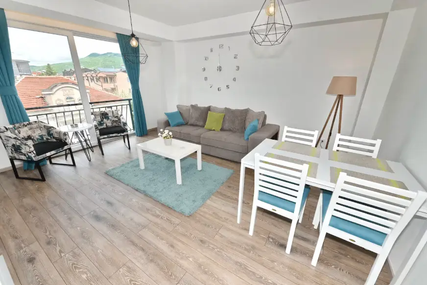 Bright, modern living room with a gray sofa, two patterned chairs, a white coffee table, a blue rug, and a dining area with a white table and four chairs, near a large window with blue curtains.
