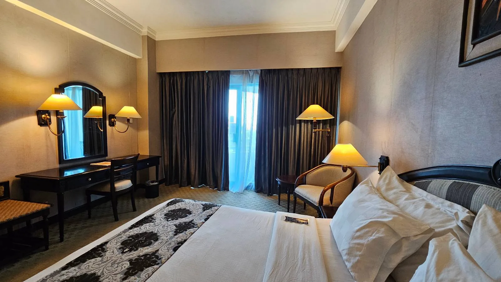 A hotel room with a neatly made bed, a patterned bedspread, and pillows. Two lamps flank the bed. A small table with a mirror and chair is in the corner. A large window with sheer and dark curtains lets in light. A cushioned chair is beside the window.