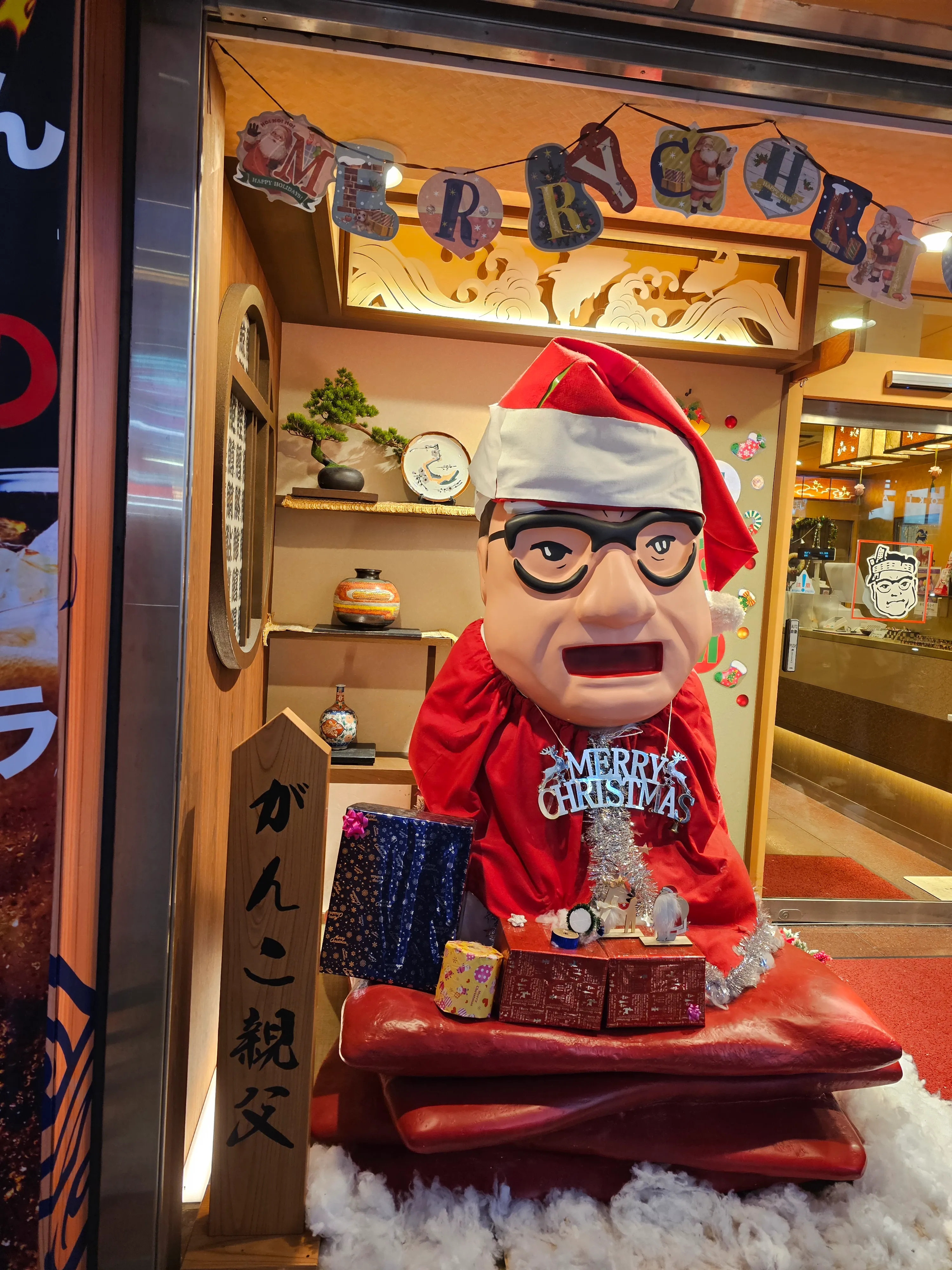 A festive shop display features a large doll with glasses and a Santa hat, wearing a red outfit and surrounded by wrapped presents. The background includes Christmas decorations and a garland spelling "Merry Christmas.