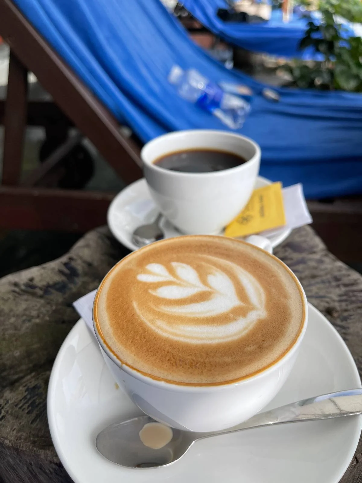 Two cups of coffee on saucers are placed on a wooden surface. The foreground cup has a latte with a leaf pattern in the foam, accompanied by a spoon. The background cup contains black coffee, with a packet of sugar, and blue chairs are partially visible.