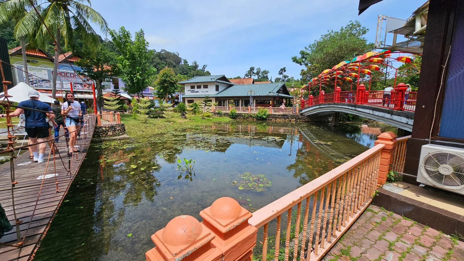 A serene park scene with a pond reflecting the sky and surrounding trees. A red bridge crosses the pond, and people stroll on paths lined with railings. Buildings with green roofs are in the background, and a palm tree adds to the tropical feel.