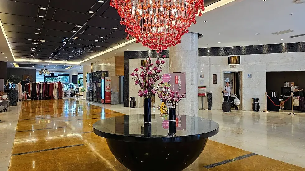 Hotel lobby with a large red chandelier, elegant flower vase on a black table. Woman with luggage exits elevator. Clothing rack in the background.