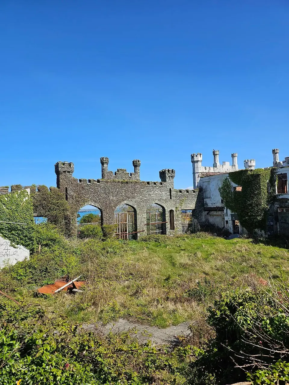 Ruins of an old stone castle with arched doorways and intact battlements under a clear blue sky. Overgrown grass and a wheelbarrow in the foreground.