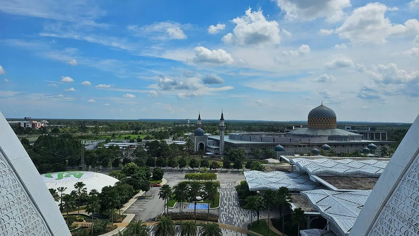 A scenic view of a sprawling urban landscape with a prominent mosque featuring a large golden dome and twin minarets. The foreground includes green trees and a geometric plaza, under a bright blue sky dotted with fluffy clouds.