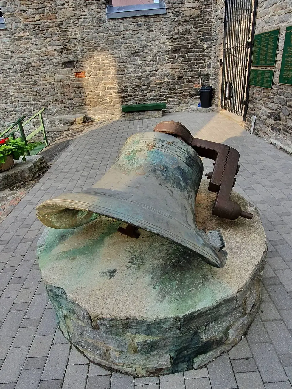 Large weathered bell on a stone pedestal against a brick wall, with green plaques and flowers in the background. Sunny setting.