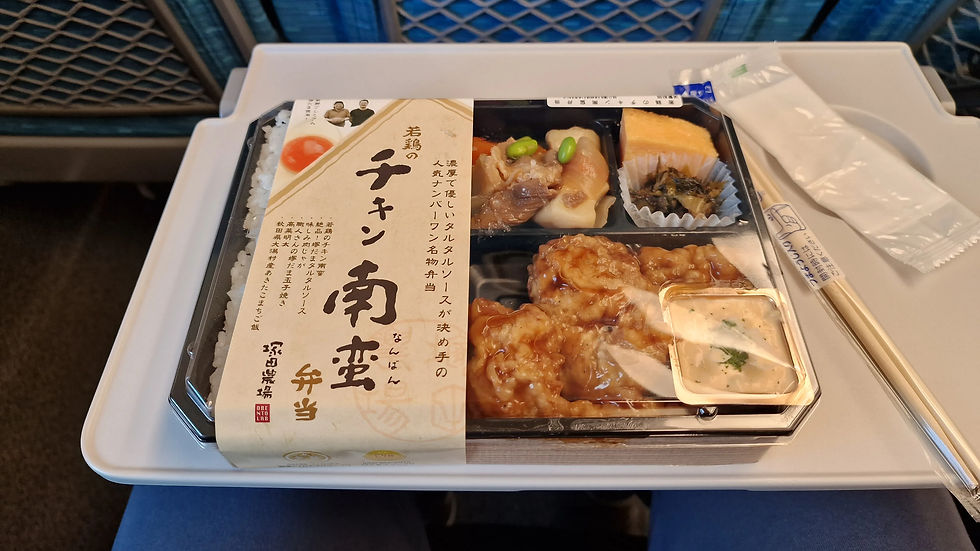 Bento box on train tray with chicken, rice, and pickles. Japanese text on packaging. Blue seat in background; chopsticks on table.