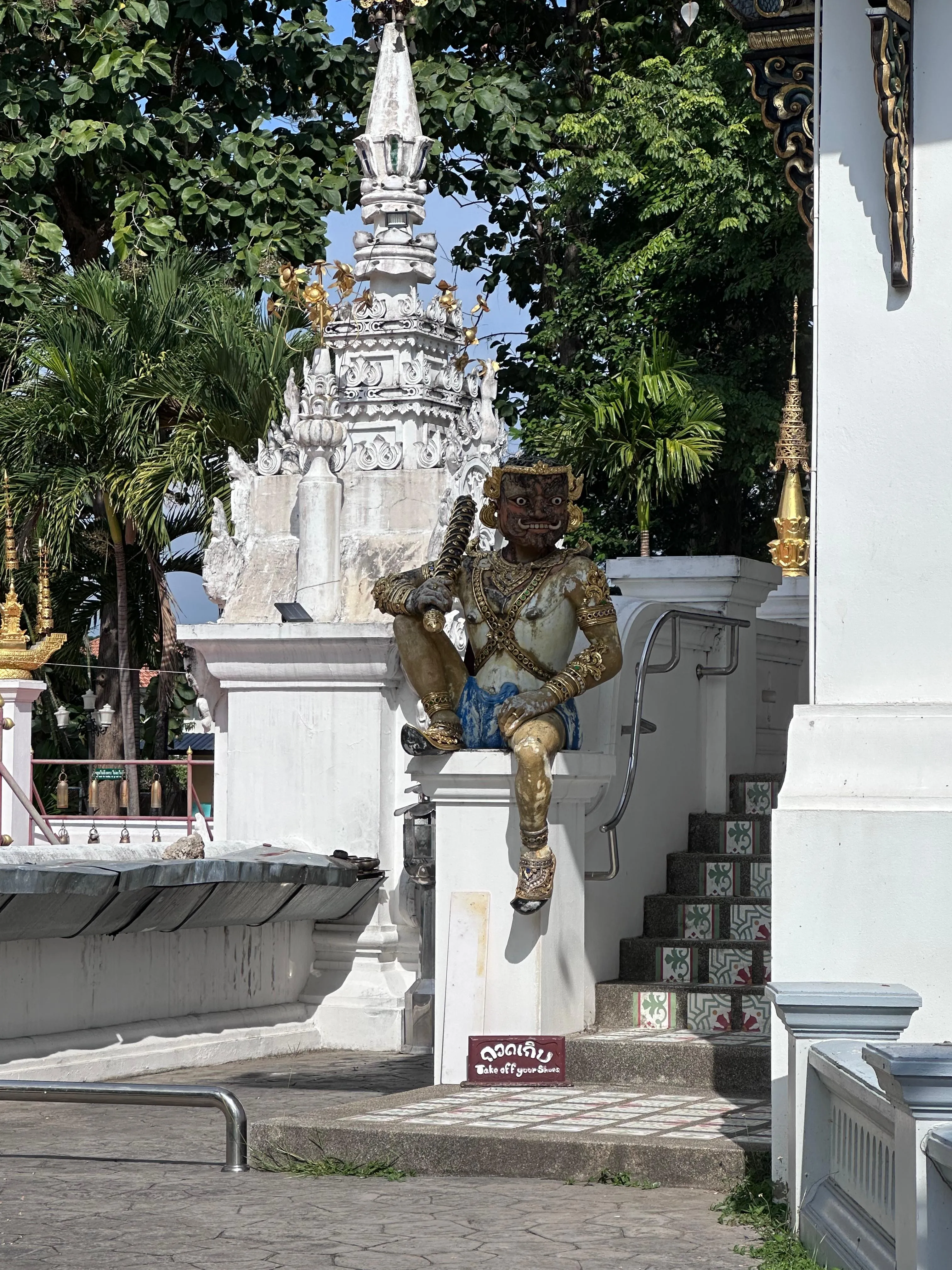 A statue of a gold-painted figure sitting on a white wall near stairs, with a white ornate structure and green trees in the background.