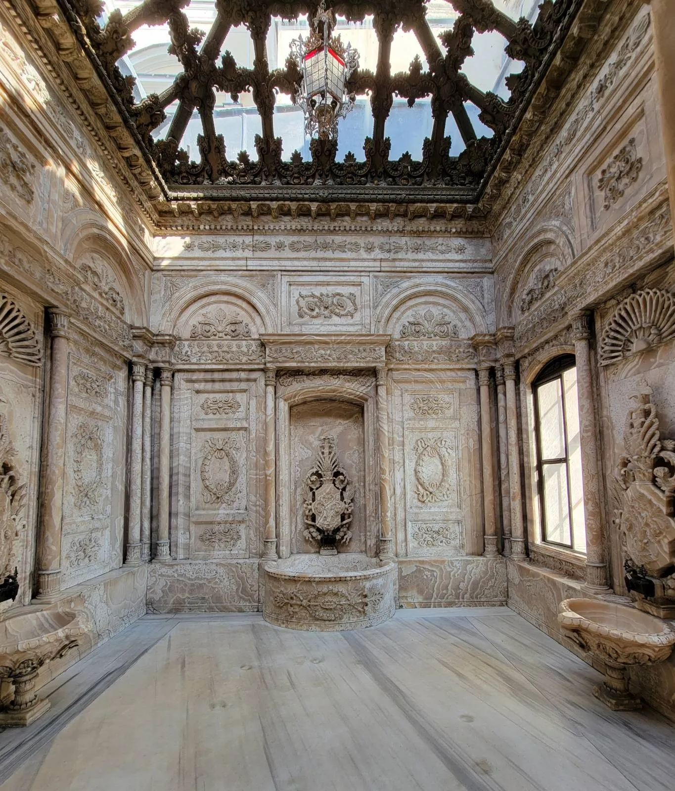 Ornate marble room with intricate carvings, arched windows, and decorative ceiling panels illuminated by natural light.