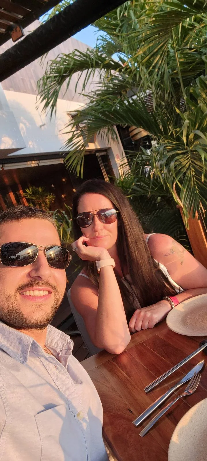 A couple wearing sunglasses is sitting at an outdoor table. The man is smiling, and the woman is resting her chin on her hand. The table is set with plates and cutlery, and there are tropical plants in the background.