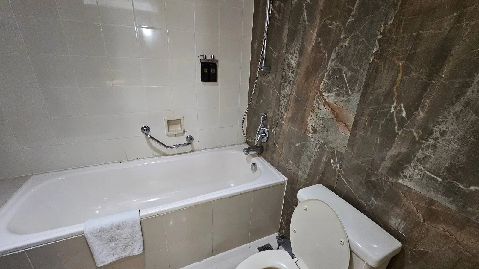 A bathroom with a white bathtub, a towel draped over its edge, and a toilet next to it. The walls are tiled, with one side featuring large, dark stone tiles.