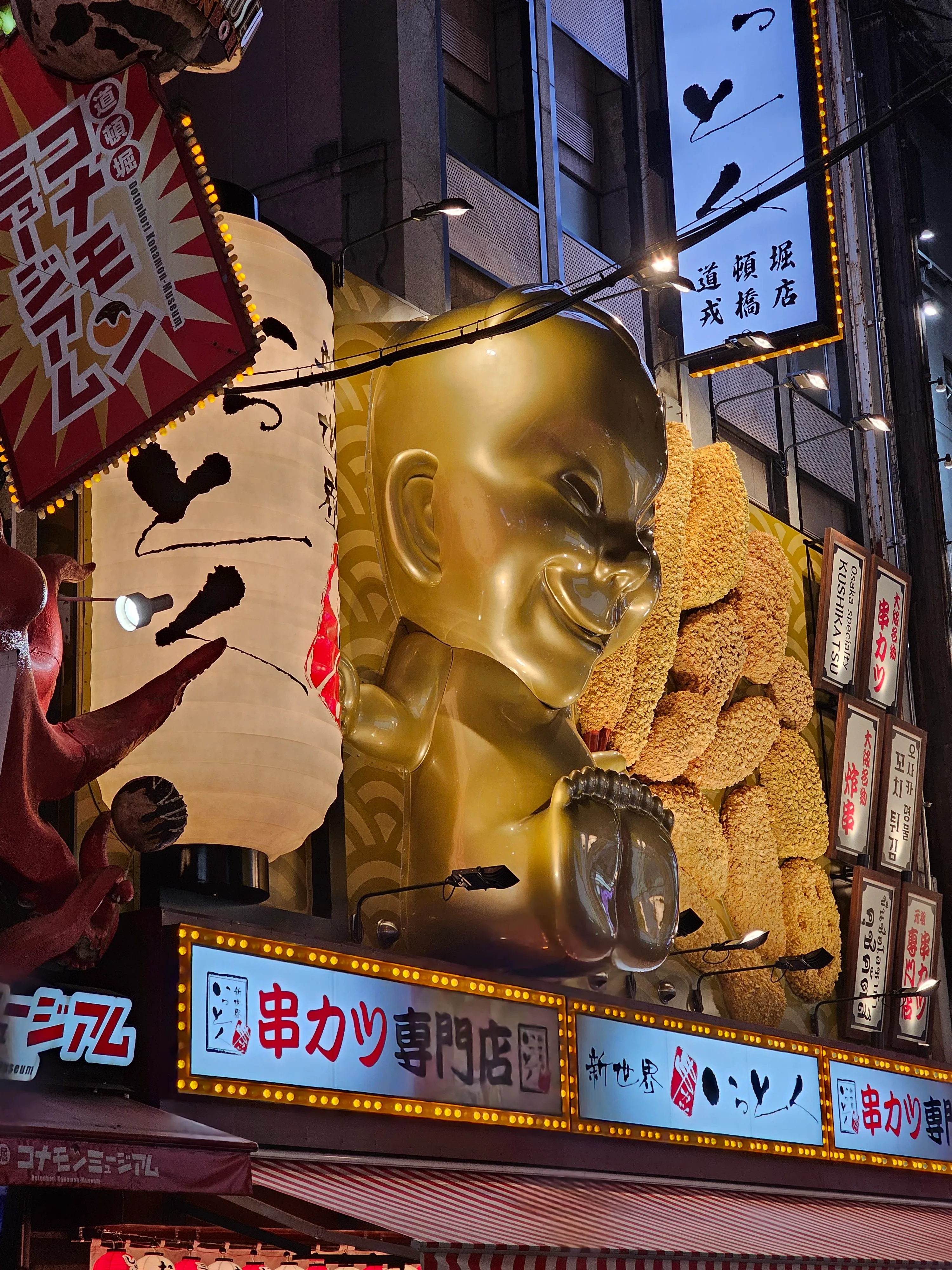 A nighttime street scene in Japan features a large golden statue of a smiling figure above a restaurant entrance, surrounded by bright neon signs and illuminated advertisements in Japanese characters.
