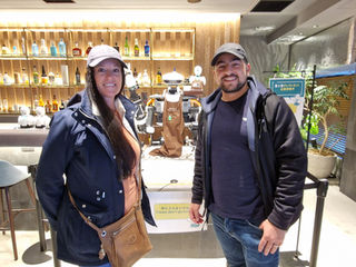 Two people stand smiling in front of an OriHime robot barista in a modern cafรฉ, highlighting the blend of technology and hospitality. The inviting atmosphere, with shelves filled with bottles, complements their casual jackets and caps, perfectly demonstrating the ease of remote working amidst health conditions.