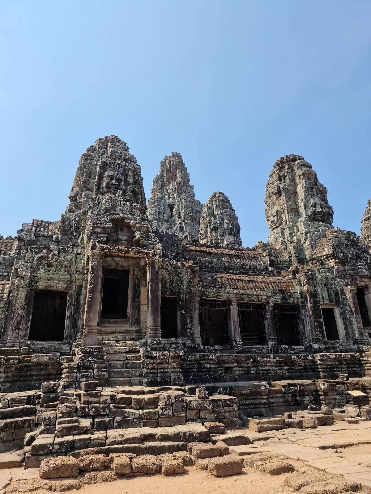 Ancient stone temple ruins with multiple towers and intricate carvings, set against a clear blue sky, likely part of Bayon Temple in Cambodia.