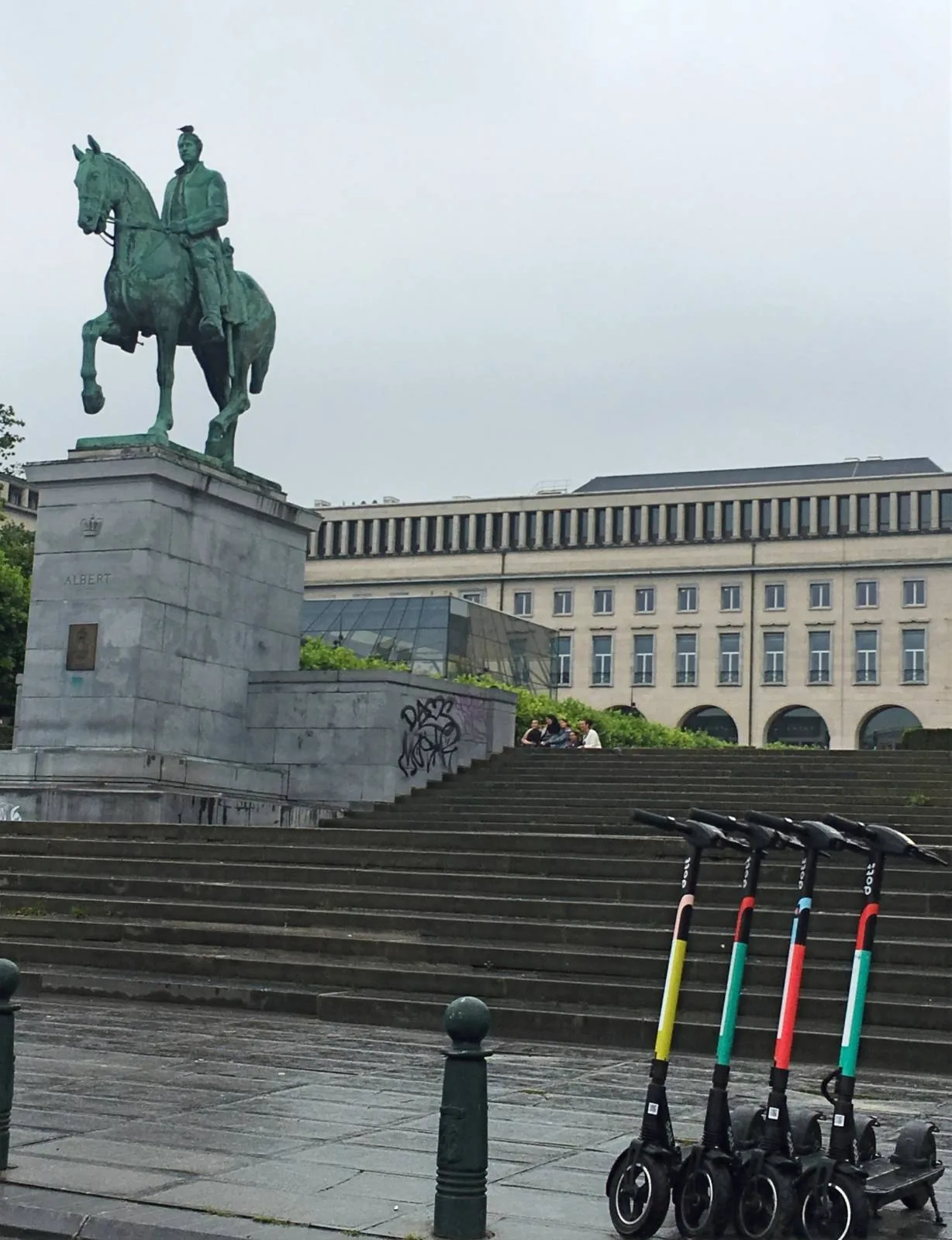 A statue of a person on horseback stands on a stone pedestal in front of a large building with multiple windows. Four electric scooters are parked in a row on the wet pavement near the statue. The sky is overcast.
