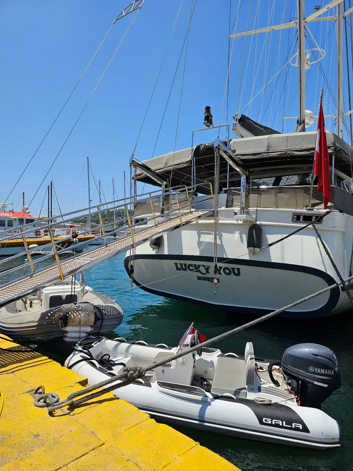 A white yacht named "Lucky You" is docked at a marina, with a small inflatable boat tied beside it. The water is blue and calm, and the sky is clear and sunny.