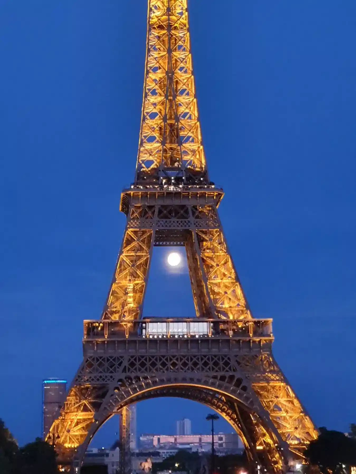 The Eiffel Tower illuminated with golden lights at night, set against a deep blue sky, with the moon visible through its structure and some city buildings at the base.