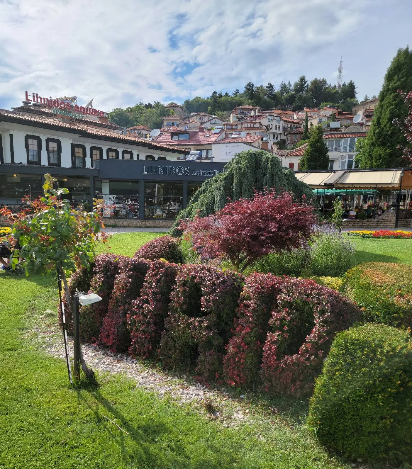 A beautifully landscaped garden featuring meticulously trimmed bushes forming the word "Ohrid." The garden is surrounded by traditional buildings with red-tiled roofs, and hills in the background under a partly cloudy sky.