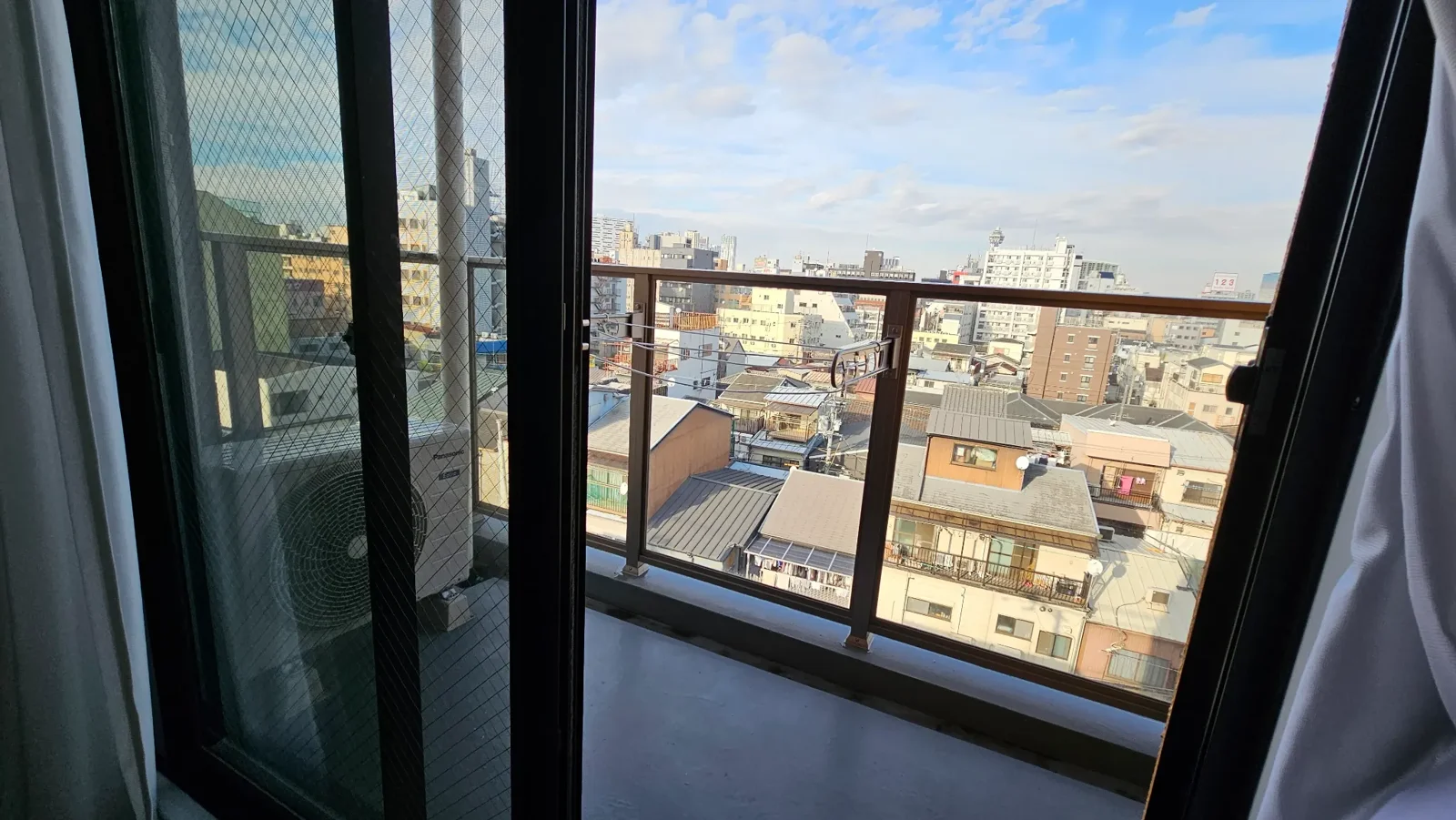 A view from inside an apartment looking out through glass sliding doors onto a balcony, with a cityscape of low-rise buildings and a blue sky with clouds in the background.