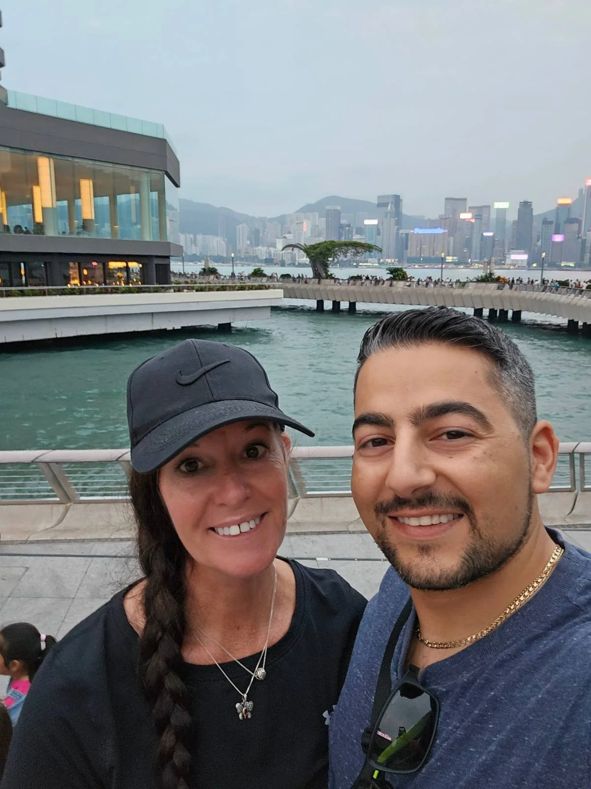 Two individuals smile for a selfie near a waterfront promenade with a cityscape and modern buildings in the background.