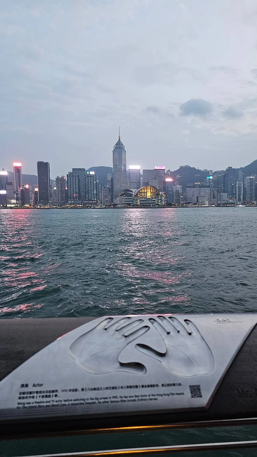 Plaque with handprint and text in the foreground; city skyline with illuminated buildings across a body of water at dusk in the background.