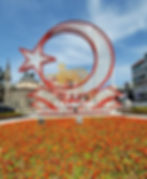 A circular monument with stars and crescent shapes, surrounded by colorful flowers, with the text "T.C. Trabzon Büyükşehir Belediyesi" in a city setting.