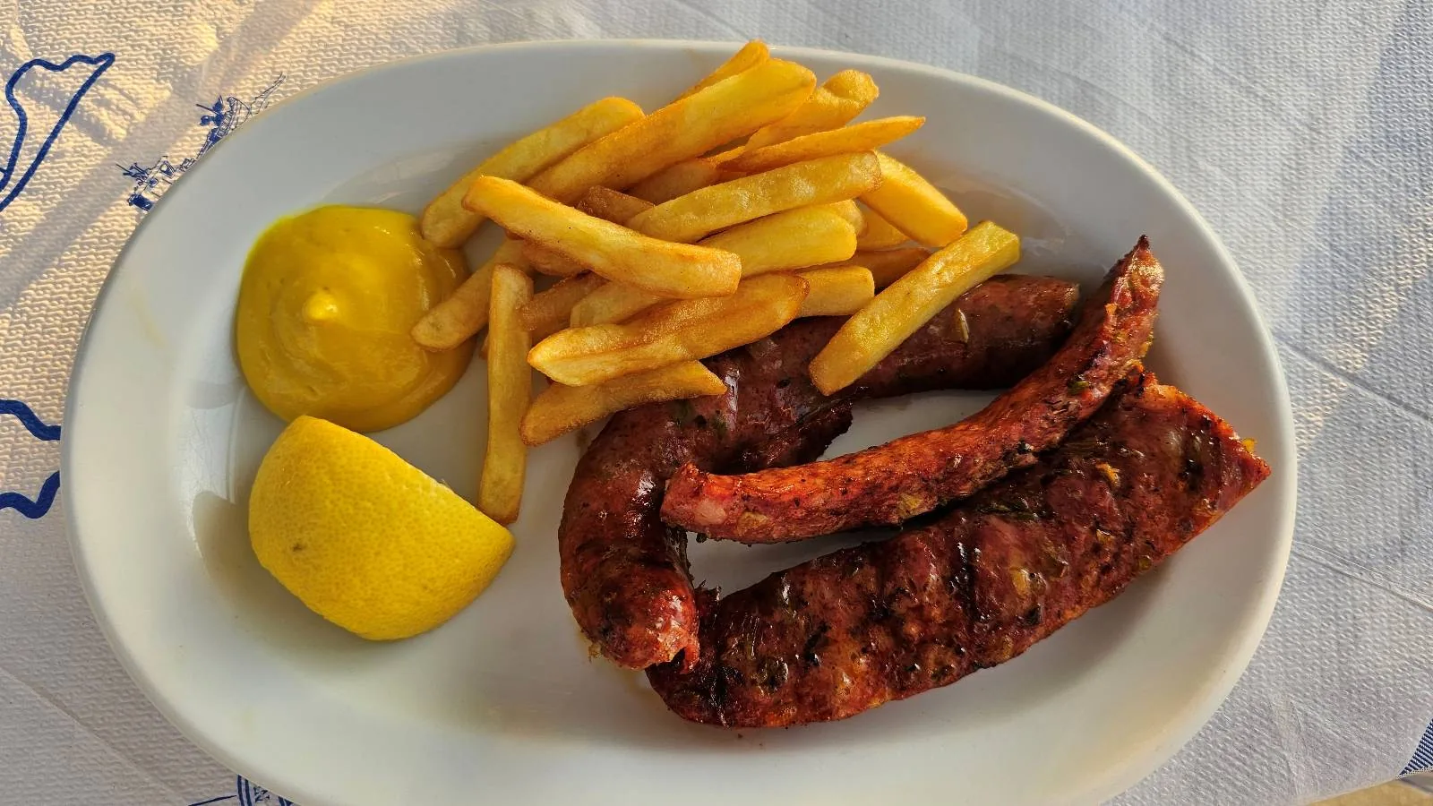 A white plate with grilled sausages, a serving of golden French fries, a dollop of yellow mustard, and a lemon wedge, set on a white tablecloth.A plate of stew featuring chunks of meat, potatoes, and carrots in a rich, brown sauce, served on a white dish with a blue and white cloth partially visible underneath.