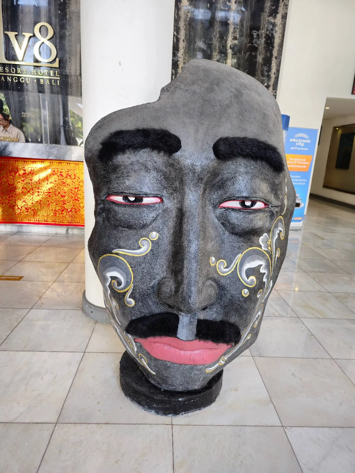 A large, sculpted face with exaggerated features like thick black eyebrows, mustache, colorful markings, and pink lips is displayed indoors on a tile floor. The face has a gray tone and intricately painted swirls on the cheeks.