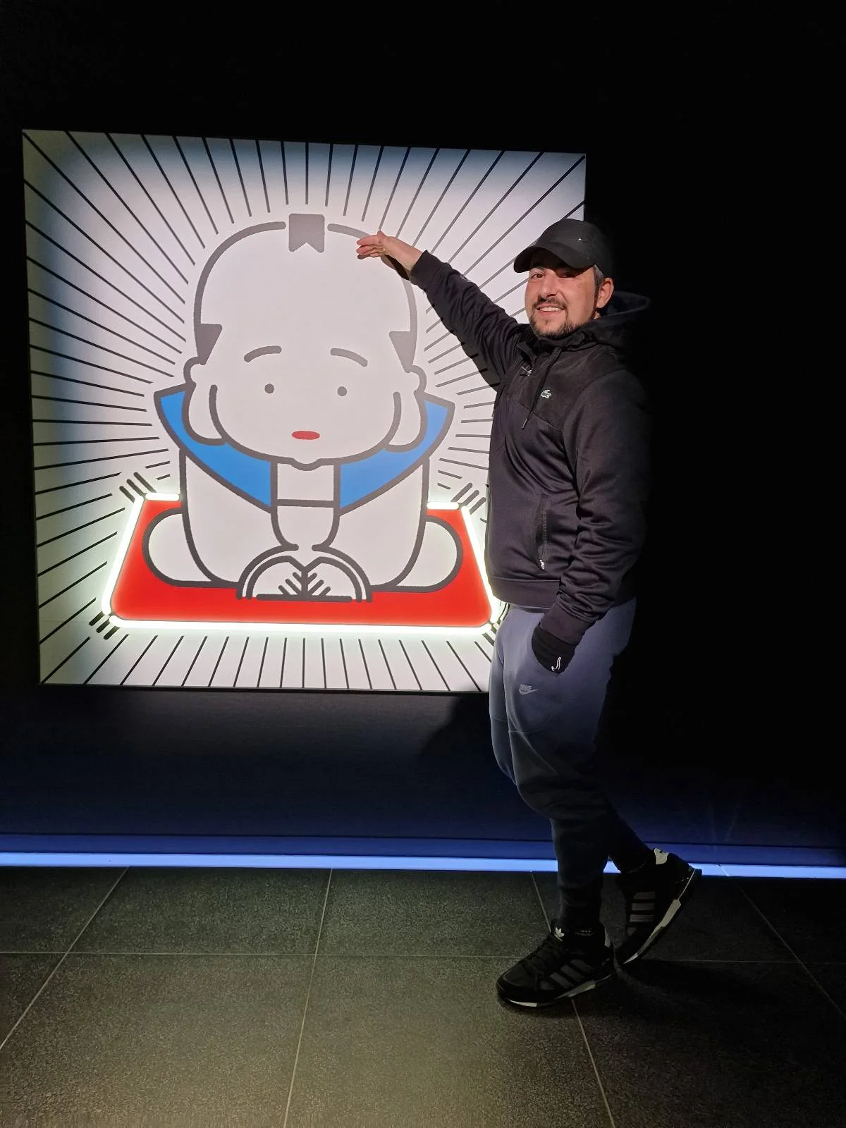 A person in dark clothing and a cap poses playfully next to a cartoon image on a wall of a child kneeling with a blue collar. The background of the image features radiating lines.