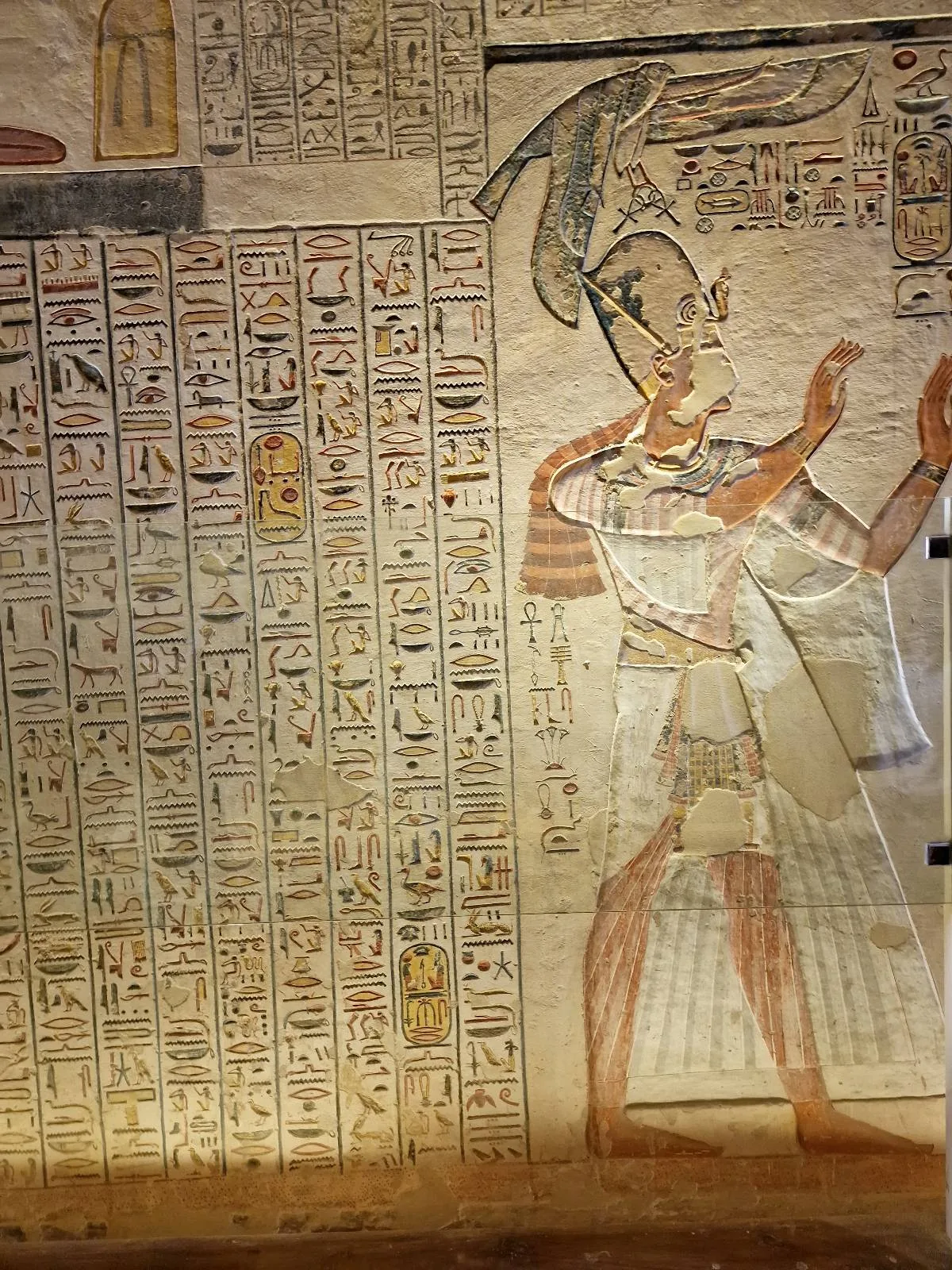 Ancient Egyptian wall painting depicting a figure with a headdress and traditional attire. Hieroglyphics cover the adjacent panels. The art is intricate and vibrant, showcasing historical and cultural significance.