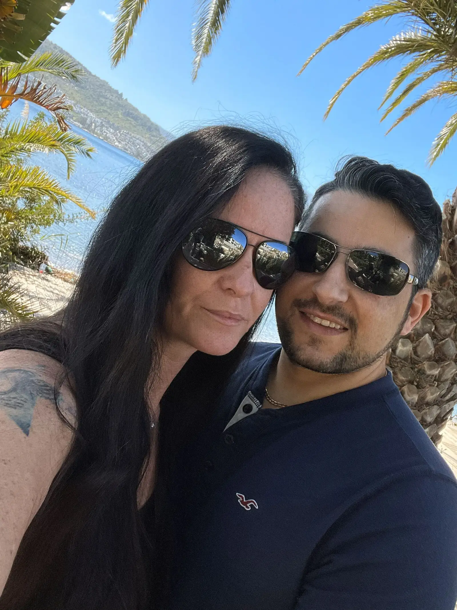 A couple wearing sunglasses poses for a selfie outdoors near palm trees and water, with blue sky and distant hills in the background.