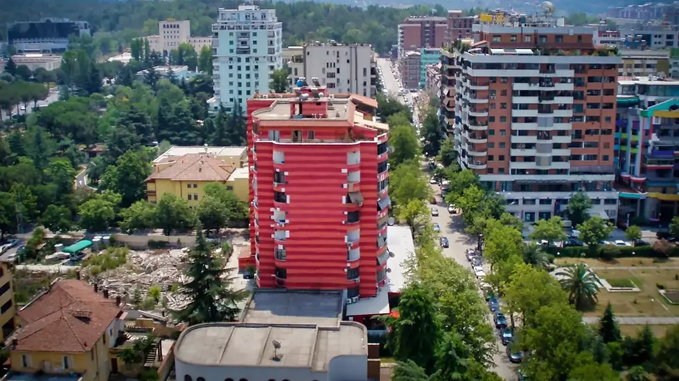 Aerial view of a cityscape with a central red striped building, surrounded by greenery and other tall buildings under a clear sky.