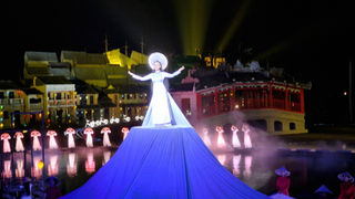 A performer in a white dress and traditional hat stands on an elevated platform. The background, reminiscent of Hoi An Memories, features illuminated traditional buildings and dancers in red and white, with reflections on water. Bright lights enhance the night scene of this vibrant cultural destination.