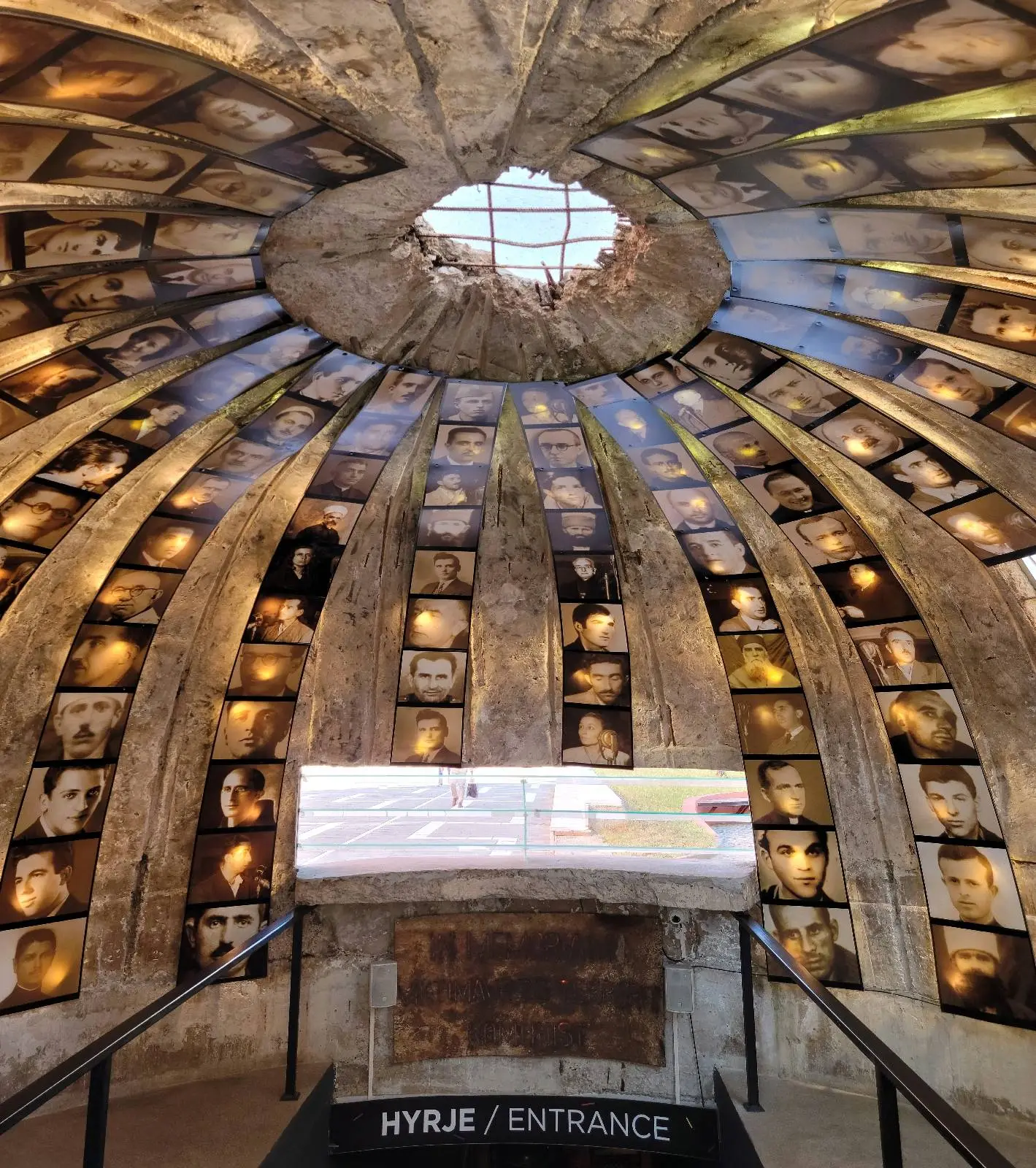 Interior view of a circular stone room with a domed ceiling, illuminated by light. The walls display numerous photographs, and a sign labeled "ENTRANCE" is visible near a railing in the foreground.