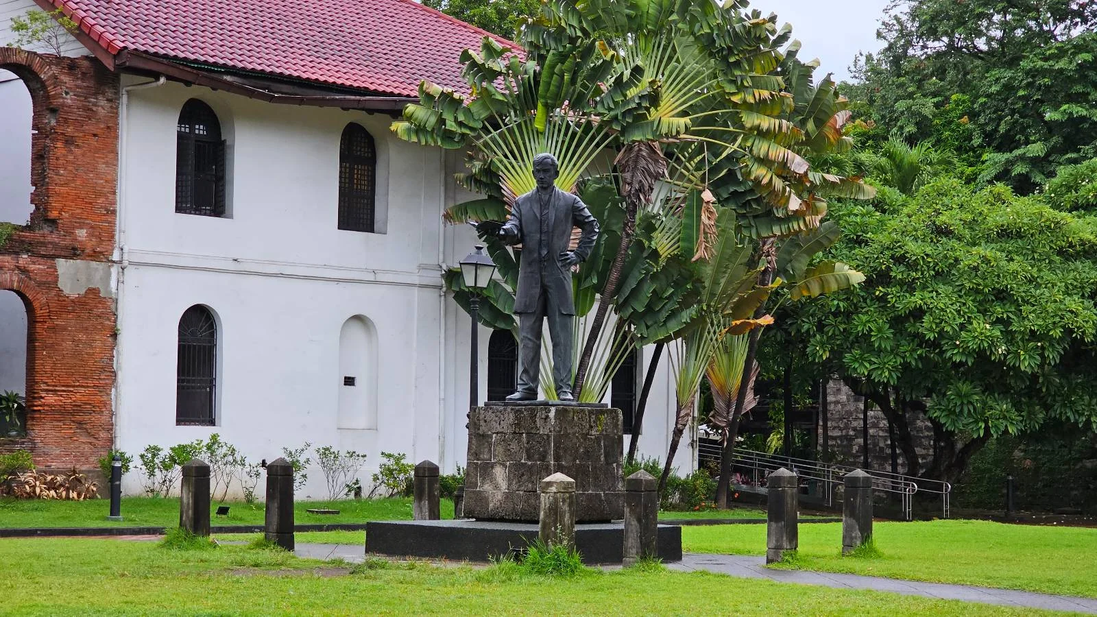 A statue of a person stands in front of a historic building with a reddish-brown roof and white walls. Lush green trees and plants surround the area, creating a serene atmosphere.