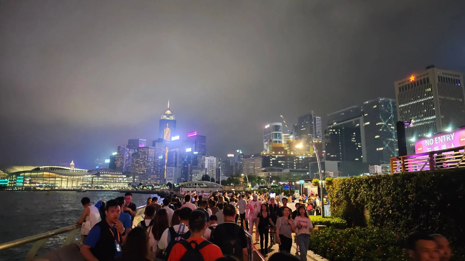 A large crowd of people are walking along a waterfront promenade at night with a skyline of illuminated skyscrapers in the background.