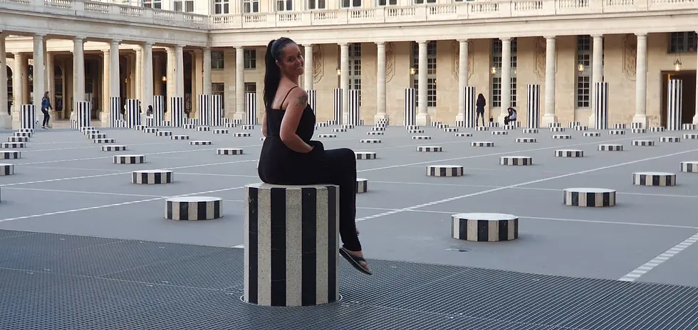 Woman in black outfit smiles while sitting on a black-and-white striped pillar. Courtyard with rows of similar pillars and classic architecture.