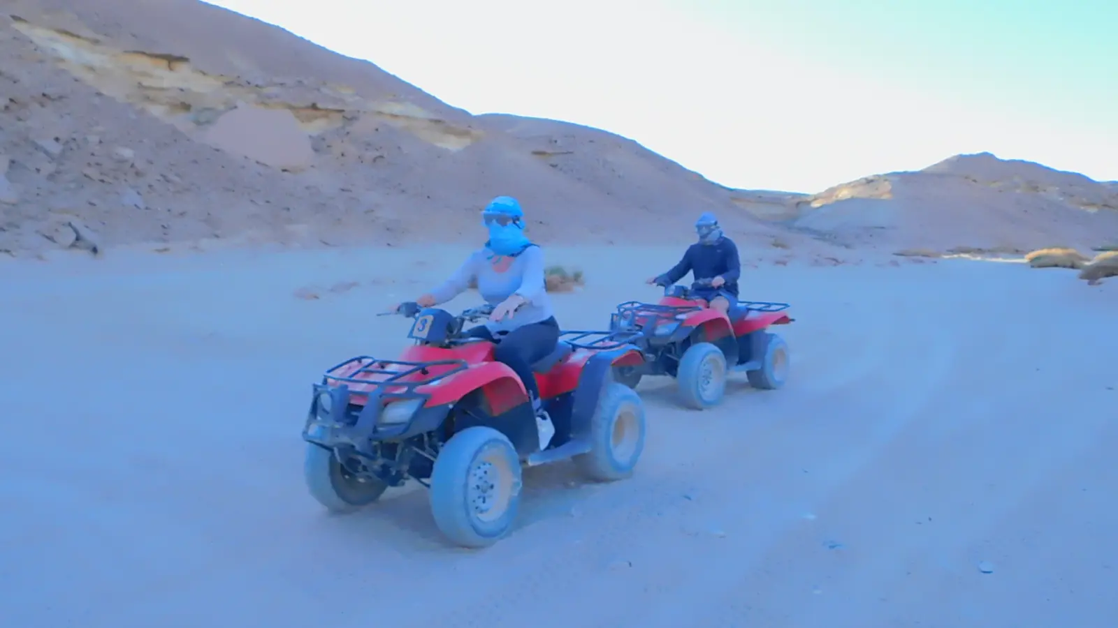 A person rides an ATV on a sandy desert landscape under a clear blue sky, followed by several others on ATVs in the background. The scene is sunlit, emphasizing the vastness of the desert.