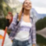 A woman is hiking, wearing a plaid shirt over a tank top that reads "BORN READY," with a tent visible in the background.